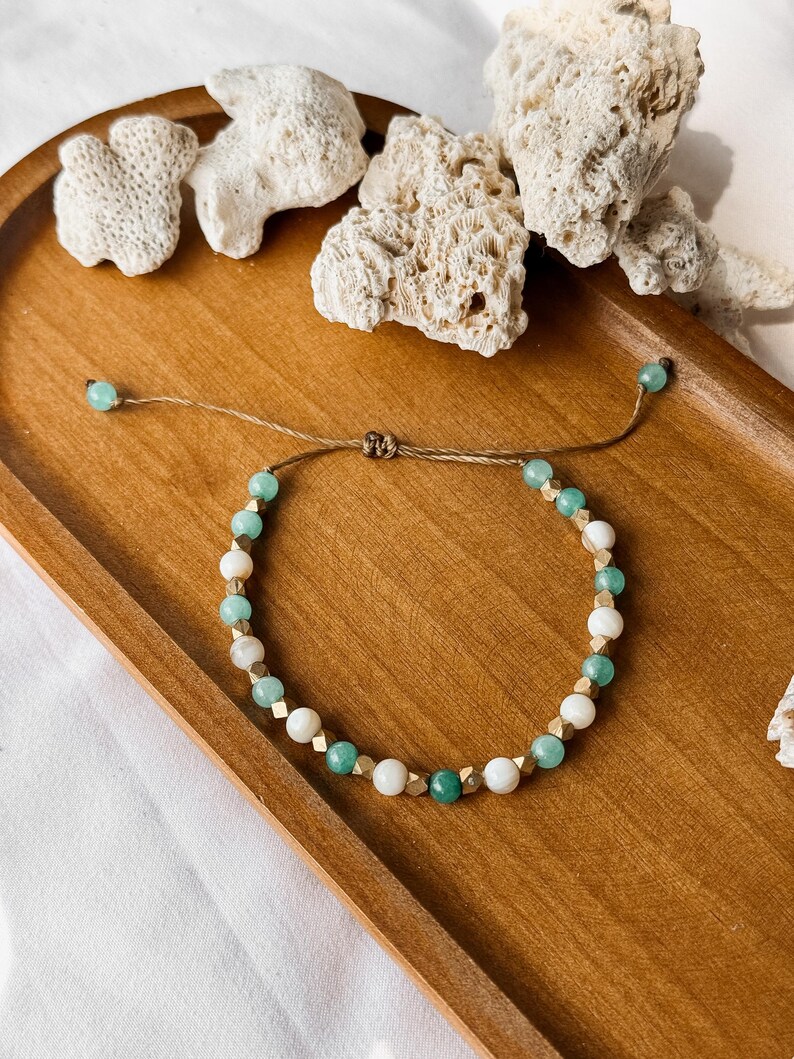 Puede incluir: Una pulsera de cuentas con una alternancia de cuentas redondas de color verde menta, cuentas blancas lisas y peque&ntilde;os espaciadores met&aacute;licos dorados. La pulsera est&aacute; ensartada en un cord&oacute;n marr&oacute;n fino con un nudo corredizo y dos cuentas verdes.