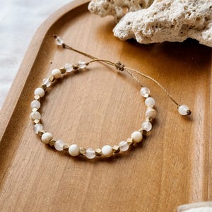 May include: A beaded bracelet featuring alternating round white beads, translucent clear beads, and small metallic gold-toned faceted cube spacers. The bracelet is strung on a thin brown cord with an adjustable sliding knot and two dangling end beads.