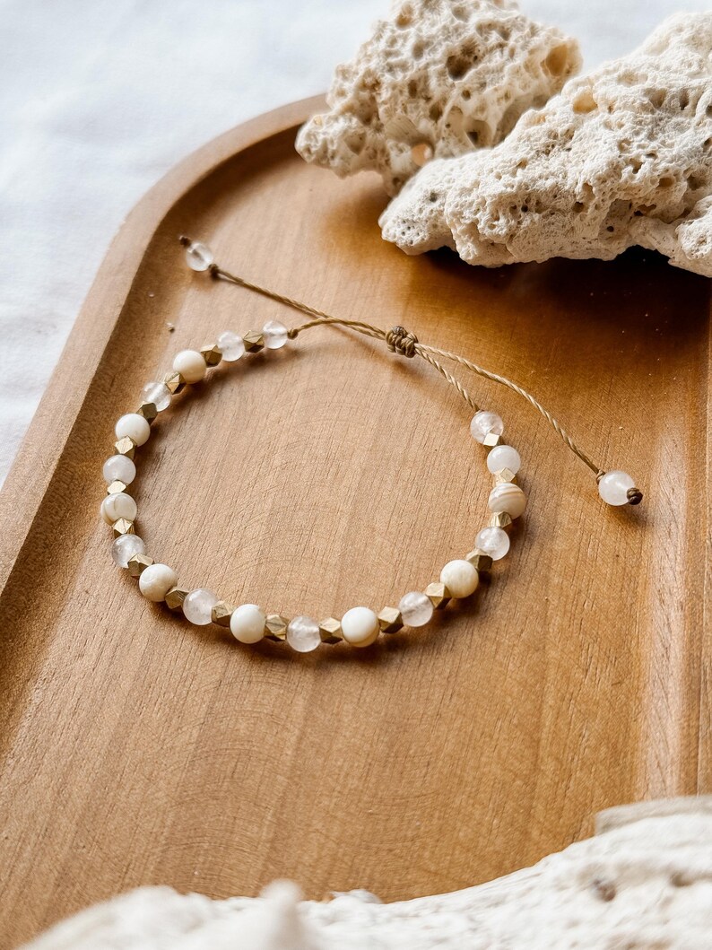 May include: A beaded bracelet featuring alternating round white beads, translucent clear beads, and small metallic gold-toned faceted cube spacers. The bracelet is strung on a thin brown cord with an adjustable sliding knot and two dangling end beads.