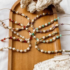 May include: A collection of beaded bracelets arranged on a wooden tray, featuring various natural stone beads in shades of red, green, purple, and white, interspersed with small metallic gold-toned spacers and strung on thin brown cord.