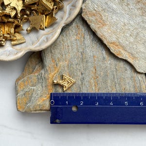 May include: A small gold arrow charm with a geometric design. The charm is about 1 inch long and is sitting on a piece of slate.