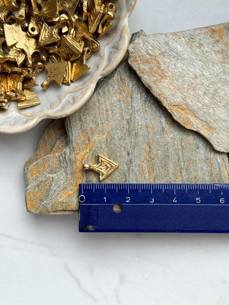 May include: A small gold arrow charm with a geometric design. The charm is about 1 inch long and is sitting on a piece of slate.