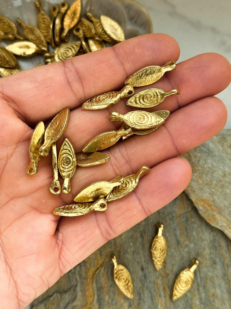 May include: A collection of gold-toned metal leaf-shaped charms with a spiral design. The charms have a hole at the top for attaching to jewelry or other crafts.