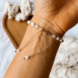 May include: A bracelet featuring small, round, translucent and opaque white beads strung on a thin, light brown cord. The design includes a sliding knot closure with two dangling cord ends, each finished with a single small, round, clear bead.