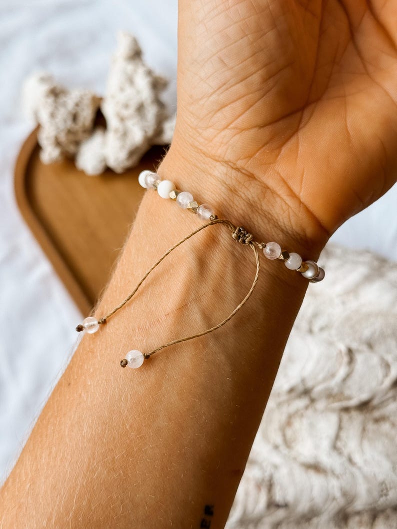 May include: A bracelet featuring small, round, translucent and opaque white beads strung on a thin, light brown cord. The design includes a sliding knot closure with two dangling cord ends, each finished with a single small, round, clear bead.