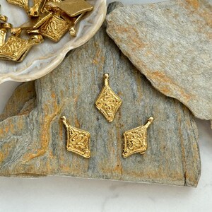 Pode incluir: Berloques de metal dourado com formato de diamante e designs ornamentados e texturizados. Alguns berloques est&atilde;o espalhados em um prato em forma de concha, enquanto outros est&atilde;o dispostos em uma superf&iacute;cie de pedra cinza. Os berloques t&ecirc;m uma argola na parte superior.