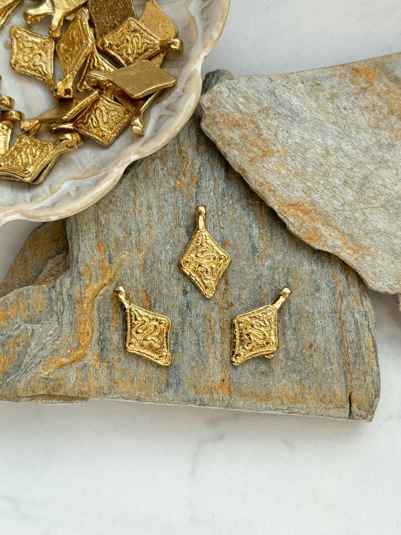 Pode incluir: Berloques de metal dourado com formato de diamante e designs ornamentados e texturizados. Alguns berloques est&atilde;o espalhados em um prato em forma de concha, enquanto outros est&atilde;o dispostos em uma superf&iacute;cie de pedra cinza. Os berloques t&ecirc;m uma argola na parte superior.