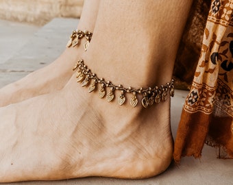 May include: A gold anklet with small leaf-shaped charms hangs on a woman's ankle. The anklet is made of a thin chain and the charms are evenly spaced.