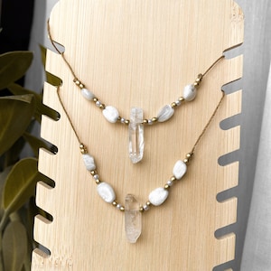 May include: Two layered necklaces on a wooden stand. Each necklace has white stone beads, small gold beads, and a clear quartz crystal pendant. The necklaces are strung on a brown cord.