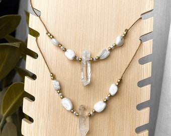 May include: Two layered necklaces displayed on a wooden stand. Each necklace features white stone beads, small gold beads, and a clear quartz crystal pendant. The necklaces are strung on a brown cord.