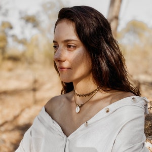May include: A woman with long brown hair wearing a white button-down shirt with the sleeves rolled up. She is wearing a gold necklace with a shell pendant. The woman is looking off to the side and has a thoughtful expression on her face.