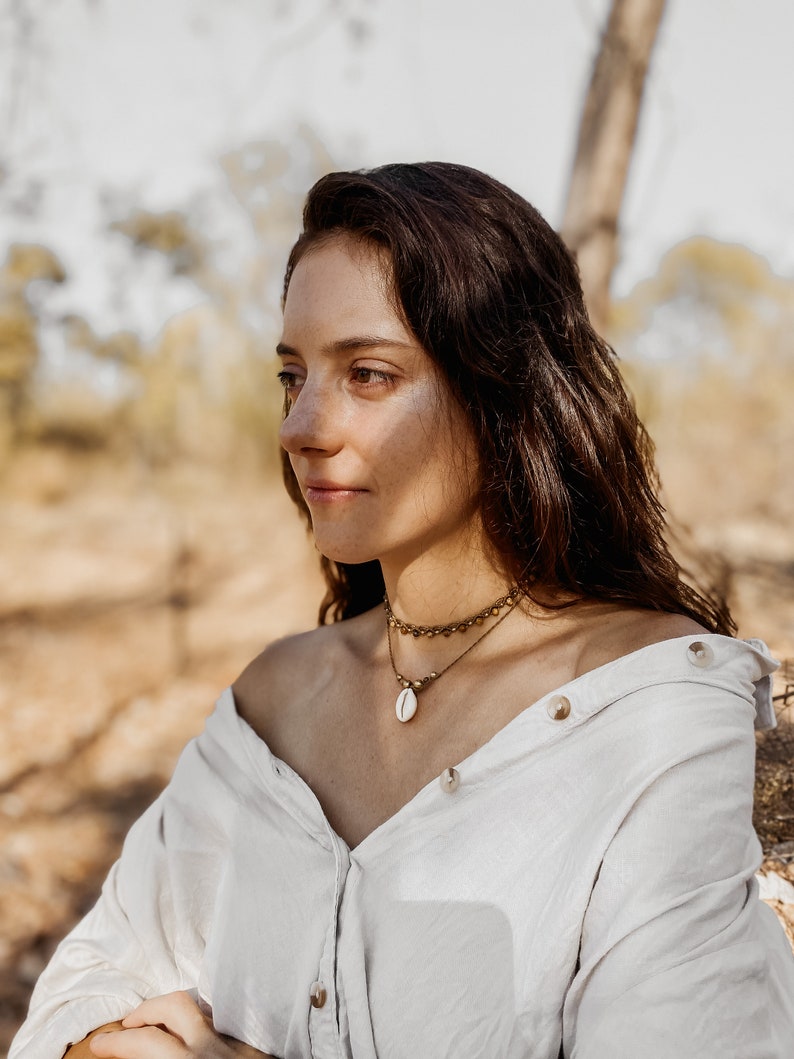 May include: A woman with long brown hair wearing a white button-down shirt with the sleeves rolled up. She is wearing a gold necklace with a shell pendant. The woman is looking off to the side and has a thoughtful expression on her face.