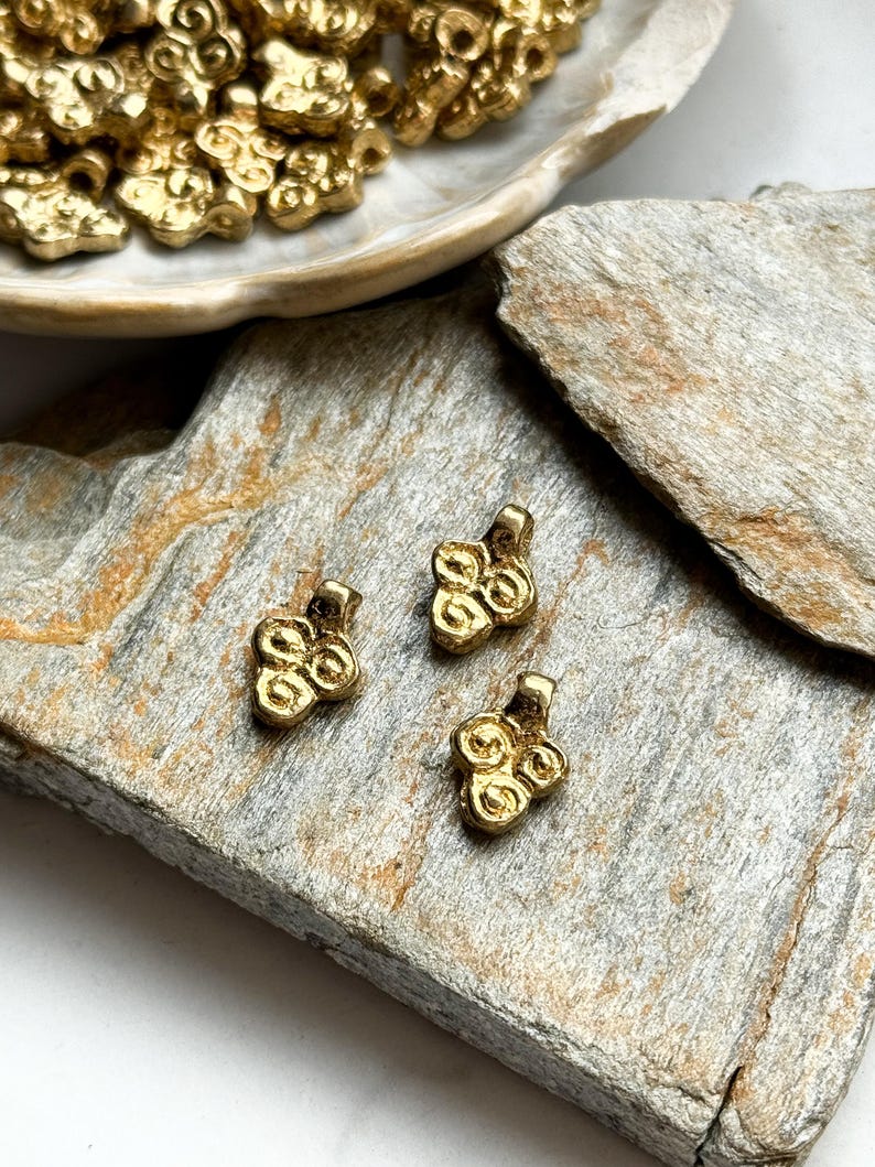May include: Three gold-colored metal charms with a swirl design. The charms are on a light brown surface.