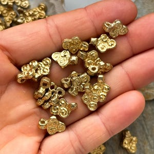 May include: A collection of gold-toned metal beads with a swirling cloud design. The beads are small and have a hole in the center for stringing.