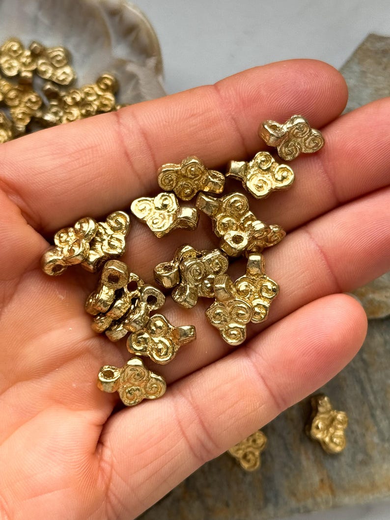 May include: A collection of gold-toned metal beads with a swirling cloud design. The beads are small and have a hole in the center for stringing.