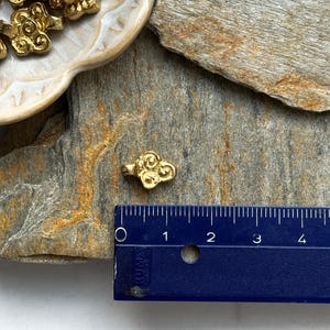 May include: Gold-colored metal charm shaped like a cloud with swirling details. The charm is about 1 inch in size and is shown next to a ruler for scale.