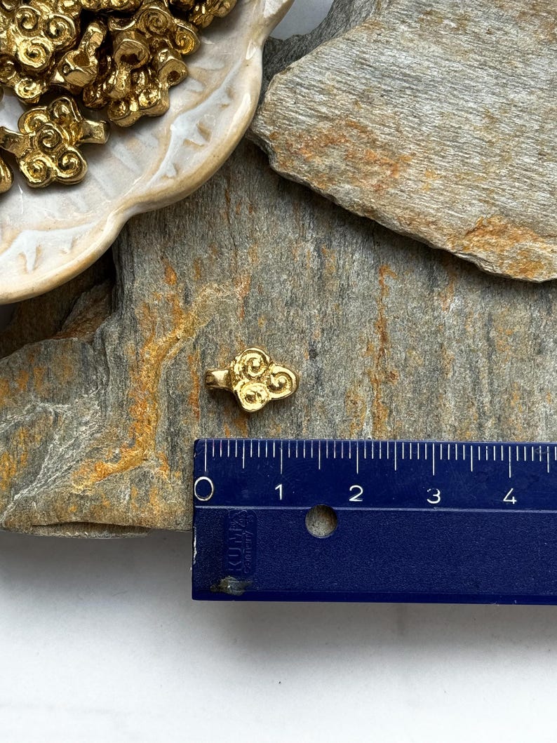 May include: Gold-colored metal charm shaped like a cloud with swirling details. The charm is about 1 inch in size and is shown next to a ruler for scale.