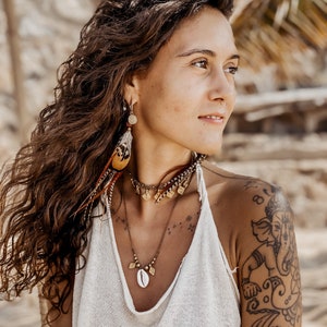 May include: A woman with long brown hair wearing a white tank top and a gold necklace with a shell pendant. She has a tattoo of an elephant on her right shoulder.