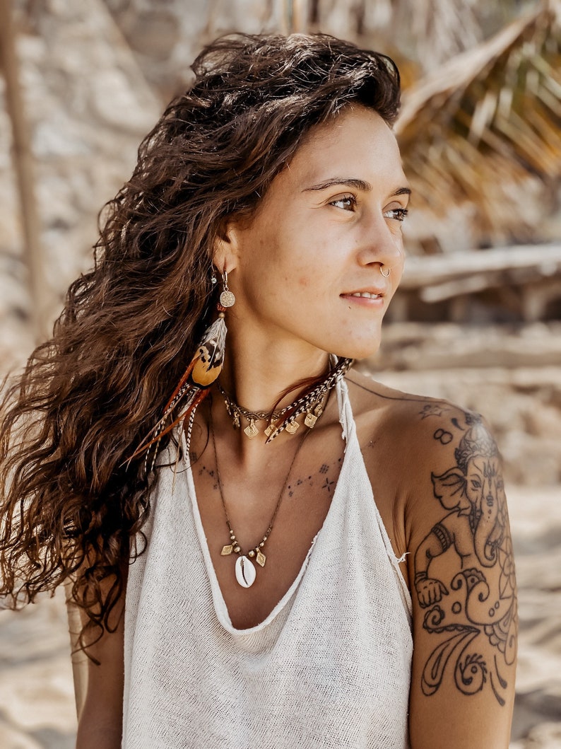 May include: A woman with long brown hair wearing a white tank top and a gold necklace with a shell pendant. She has a tattoo of an elephant on her right shoulder.