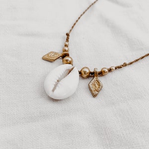 May include: A gold necklace with a white cowrie shell pendant and two gold diamond-shaped charms. The necklace is made of brown cord and has gold beads.