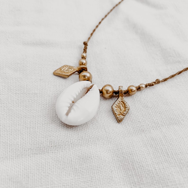 May include: A gold necklace with a white cowrie shell pendant and two gold diamond-shaped charms. The necklace is made of brown cord and has gold beads.