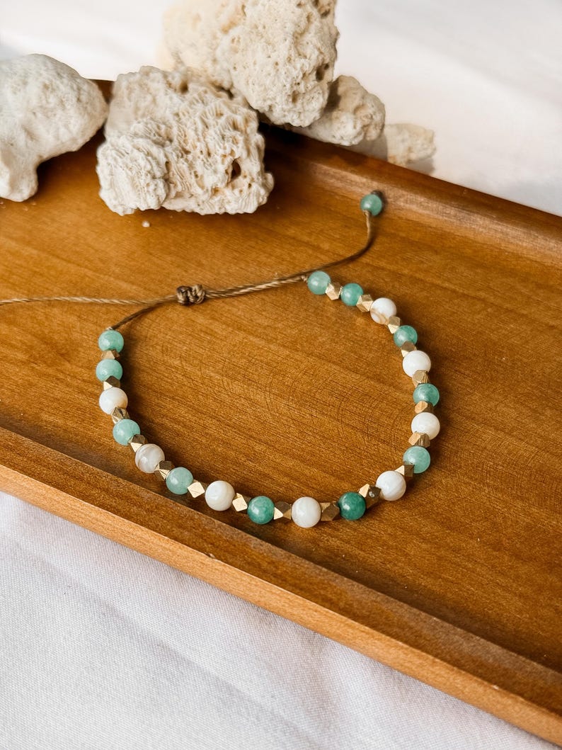 Puede incluir: Una pulsera de cuentas que alterna cuentas redondas de color verde menta, cuentas blancas lisas y peque&ntilde;os espaciadores met&aacute;licos facetados en tono dorado. La pulsera est&aacute; hecha con un cord&oacute;n marr&oacute;n fino y un cierre de nudo corredizo.