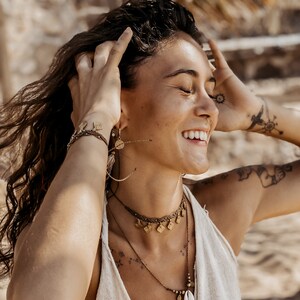 May include: A woman with long brown hair wearing a white tank top and multiple gold necklaces with charms. She is smiling and has her hands in her hair.