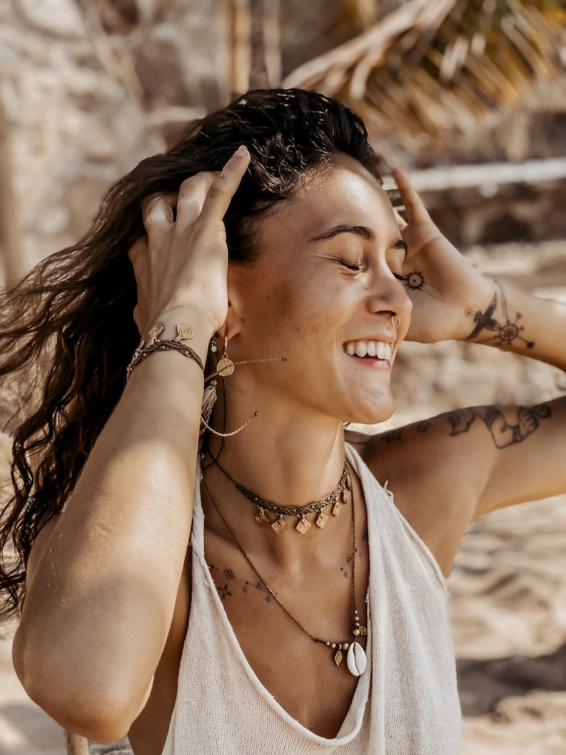 May include: A woman with long brown hair wearing a white tank top and multiple gold necklaces with charms. She is smiling and has her hands in her hair.