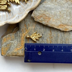 以下が含まれることがあります： シンプルな葉の形をした金の葉のチャーム。チャームの長さは約2.5cmで、サイズ比較のために定規の隣に置かれています。