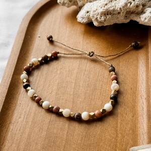 May include: A beaded bracelet featuring alternating round white, brown, and terracotta-colored beads interspersed with small, faceted metallic gold-toned cubes. The bracelet is strung on a thin brown cord with an adjustable sliding knot closure.