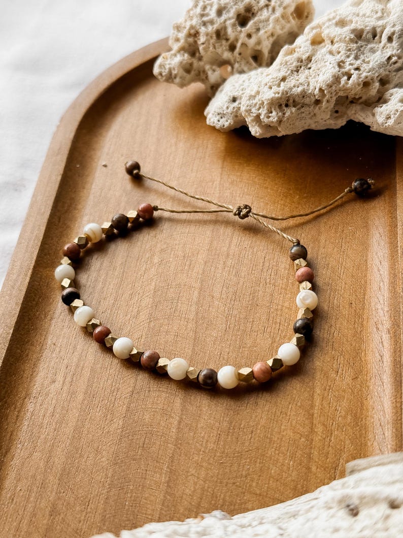 May include: A beaded bracelet featuring alternating round white, brown, and terracotta-colored beads interspersed with small, faceted metallic gold-toned cubes. The bracelet is strung on a thin brown cord with an adjustable sliding knot closure.