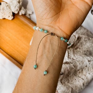 Puede incluir: Una pulsera de cuentas con cuentas esf&eacute;ricas alternas en verde claro y blanco con peque&ntilde;os detalles en tono dorado. La pulsera tiene un cierre de cord&oacute;n marr&oacute;n con dos hilos colgantes, cada uno terminado con una cuenta verde claro al final.