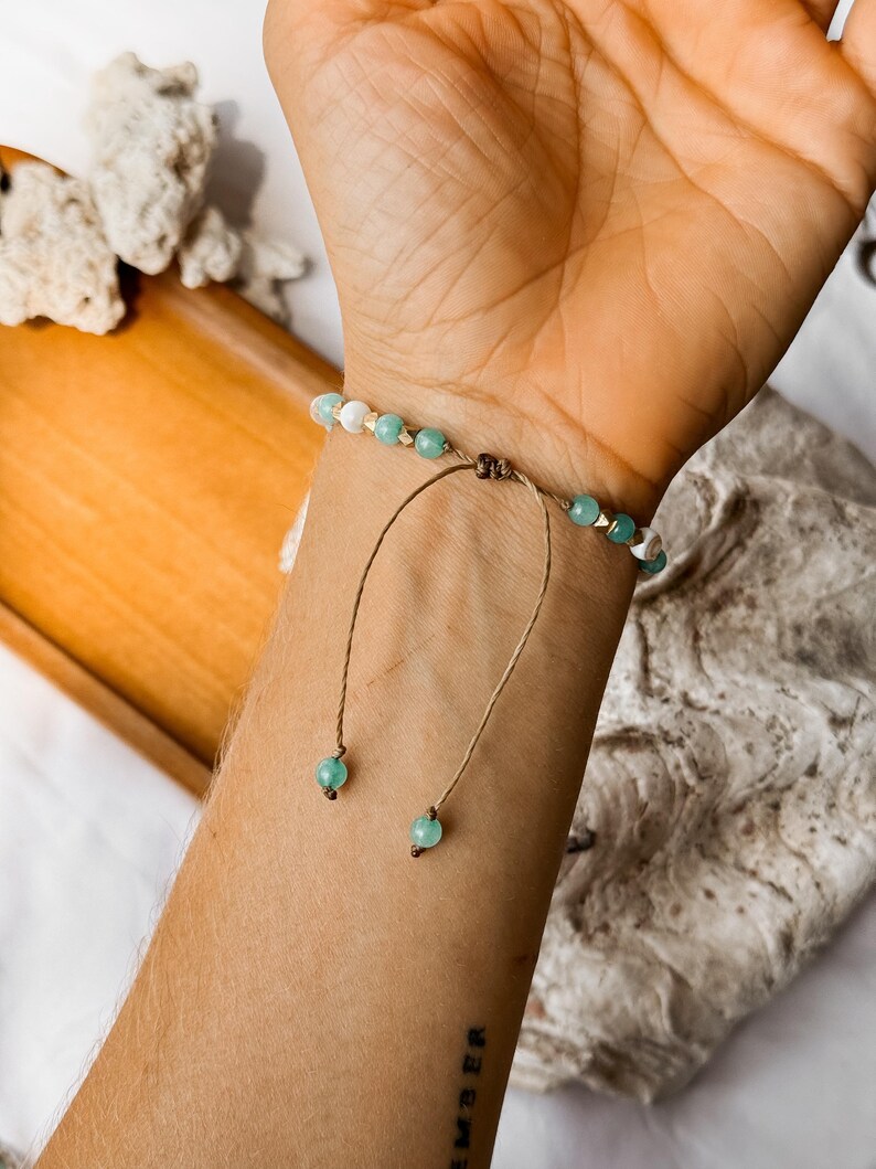 Puede incluir: Una pulsera de cuentas con cuentas esf&eacute;ricas alternas en verde claro y blanco con peque&ntilde;os detalles en tono dorado. La pulsera tiene un cierre de cord&oacute;n marr&oacute;n con dos hilos colgantes, cada uno terminado con una cuenta verde claro al final.