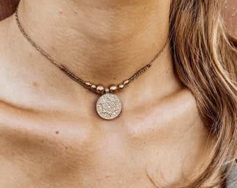 May include: A gold choker necklace with a round pendant featuring an intricate design. The necklace is made of brown cord and has small gold beads.