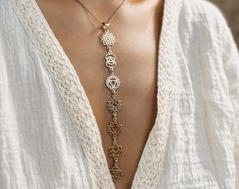May include: A gold-colored necklace featuring seven chakra symbols, suspended vertically. The pendant is worn against a white garment with lace trim. The necklace is a delicate piece of jewelry.