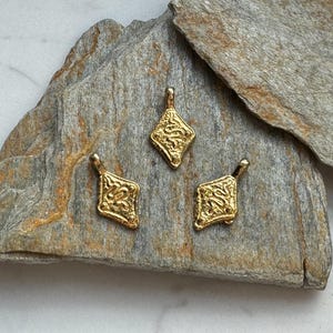 Pode incluir: Tr&ecirc;s pingentes dourados com formato de diamante e designs ornamentados e texturizados. Cada pingente tem uma pequena argola na parte superior para pendurar. Os pingentes s&atilde;o exibidos em uma superf&iacute;cie de pedra cinza texturizada.
