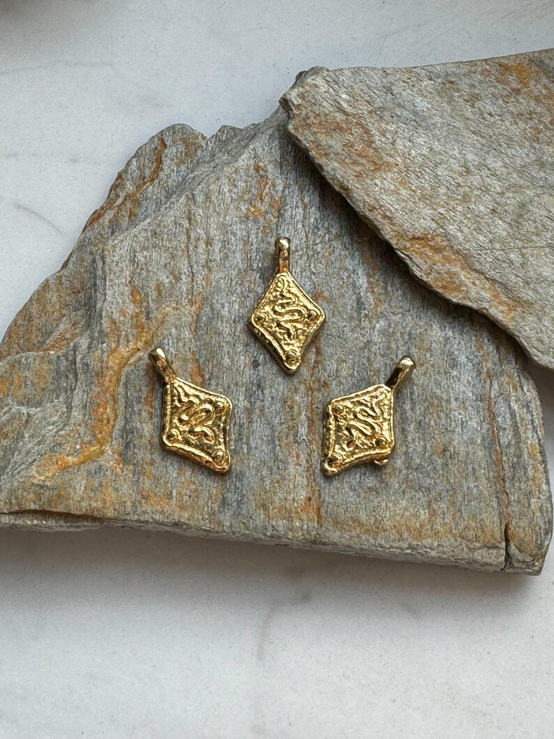 Pode incluir: Tr&ecirc;s pingentes dourados com formato de diamante e designs ornamentados e texturizados. Cada pingente tem uma pequena argola na parte superior para pendurar. Os pingentes s&atilde;o exibidos em uma superf&iacute;cie de pedra cinza texturizada.