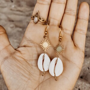 May include: A pair of gold earrings with a sun charm and a white shell pendant. The earrings are on a hand with a gold ring.