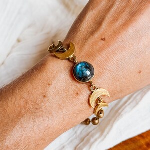 May include: A bracelet featuring a central round cabochon of iridescent blue labradorite stone, flanked by multiple brass-toned crescent moon charms and small spherical beads, connected by metal links in a chain-style arrangement.