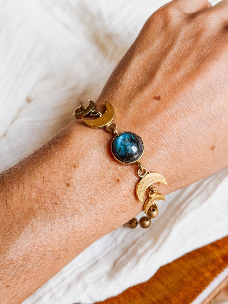 May include: A bracelet featuring a central round cabochon of iridescent blue labradorite stone, flanked by multiple brass-toned crescent moon charms and small spherical beads, connected by metal links in a chain-style arrangement.