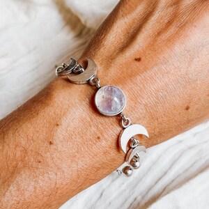 May include: A silver-toned bracelet featuring a central round iridescent stone flanked by multiple crescent moon charms. The chain links connect the moon motifs and the circular gemstone, creating a celestial-themed piece of jewelry.