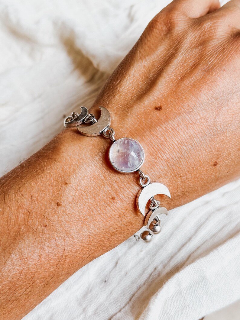 May include: A silver-toned bracelet featuring a central round iridescent stone flanked by multiple crescent moon charms. The chain links connect the moon motifs and the circular gemstone, creating a celestial-themed piece of jewelry.