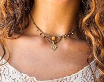 May include: A brown macrame choker necklace featuring a central round tan stone bead, a small diamond-shaped brass pendant with etched geometric lines, and a series of tiny dangling brass accents along the woven cord band.