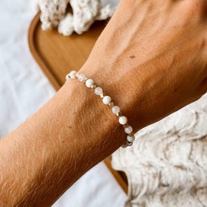 May include: A beaded bracelet featuring a repeating pattern of small, round, opaque white beads and translucent, faceted clear beads, interspersed with tiny, metallic gold-toned spacer beads, worn around a wrist.