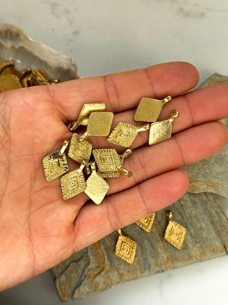 Puede incluir: Una colecci&oacute;n de dijes de metal dorado en forma de diamante con intrincados dise&ntilde;os geom&eacute;tricos. Cada dije tiene un peque&ntilde;o bucle en la parte superior para sujetarlo a joyas u otras manualidades.