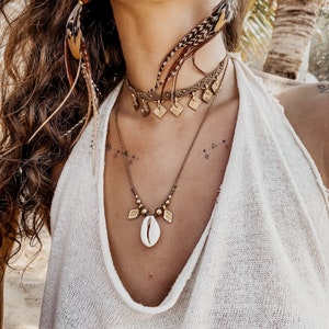 May include: A woman wearing a white crochet top with a gold shell necklace and feather earrings. The necklace has a small shell pendant and gold beads.