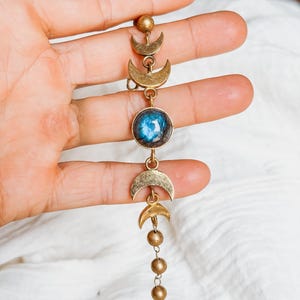 May include: A gold-toned metal bracelet featuring a series of crescent moon charms and a central circular stone with a blue iridescent sheen. The design includes small spherical beads and delicate chain links, creating a linear arrangement of celestial-inspired elements.