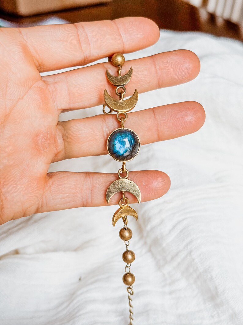 May include: A gold-toned metal bracelet featuring a series of crescent moon charms and a central circular stone with a blue iridescent sheen. The design includes small spherical beads and delicate chain links, creating a linear arrangement of celestial-inspired elements.