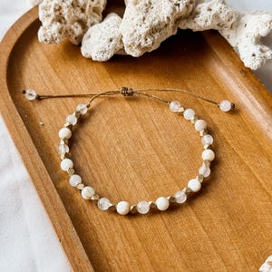 May include: A delicate adjustable bracelet featuring a repeating pattern of round white beads, translucent clear beads, and small metallic gold-toned faceted spacers. The bracelet is strung on a thin brown cord with a sliding knot closure mechanism.