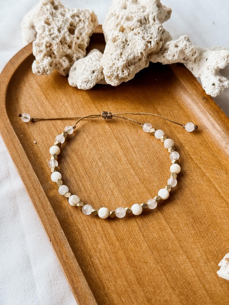 May include: A delicate adjustable bracelet featuring a repeating pattern of round white beads, translucent clear beads, and small metallic gold-toned faceted spacers. The bracelet is strung on a thin brown cord with a sliding knot closure mechanism.